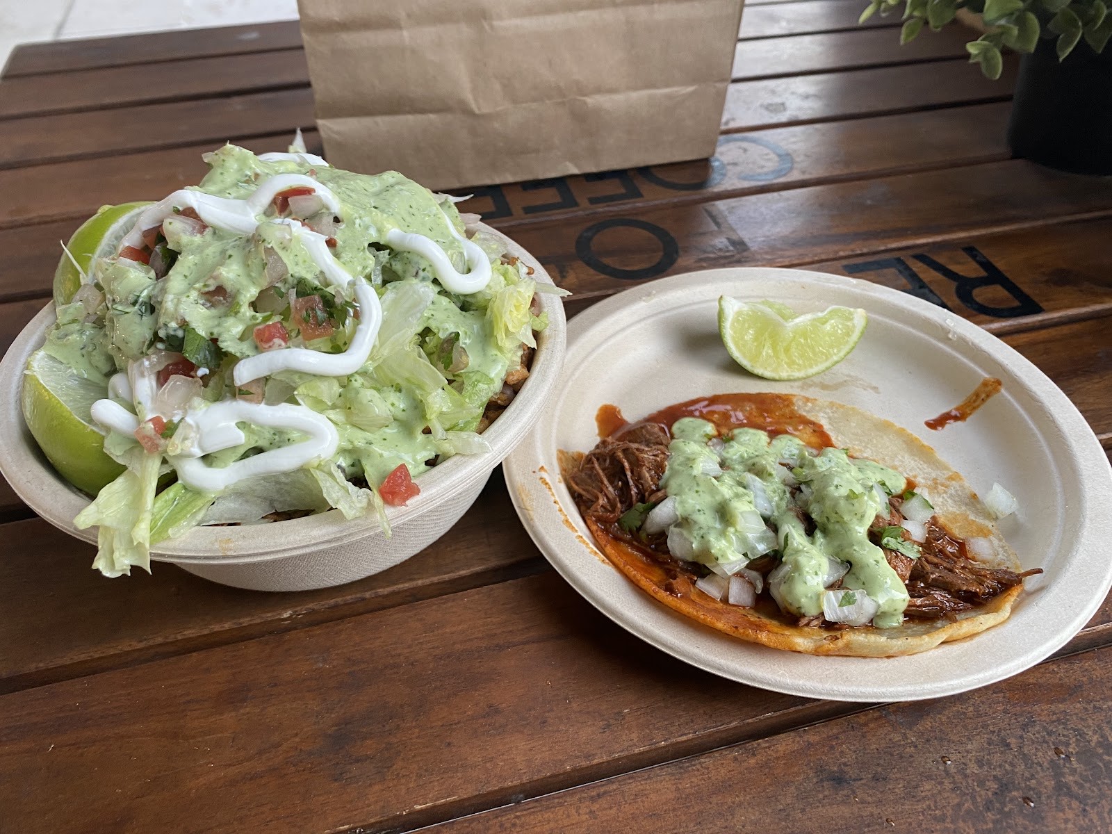 A birria-style beef taco in red consommé topped with creamy green salsa and onion, beside a loaded bowl