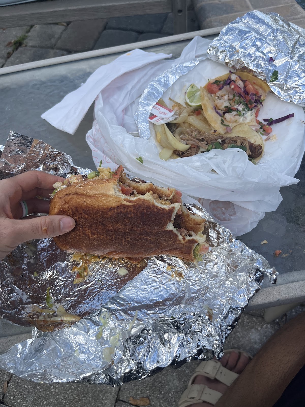 A toasted Torta de Al Pastor sandwich on a crusty roll held in hand, with fish tacos in the background